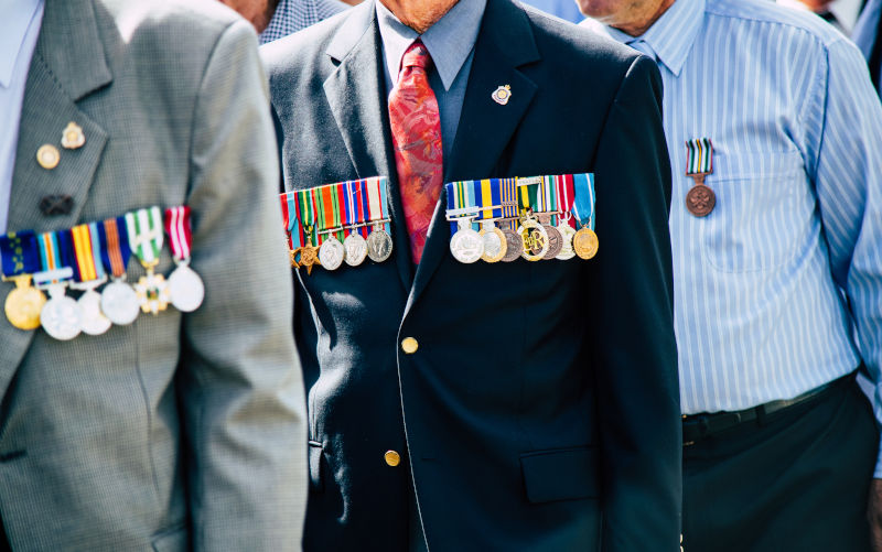 A formed body of old military veterans march past the crowd during the Cooroy-Pomona ANZAC Day parade. The man in the foreground proudly wears the golden Order of Australia Medal (OAM) along with campaign medals from The Vietnam War, the Infantry Combat Badge (ICB) and general Defence service medals.