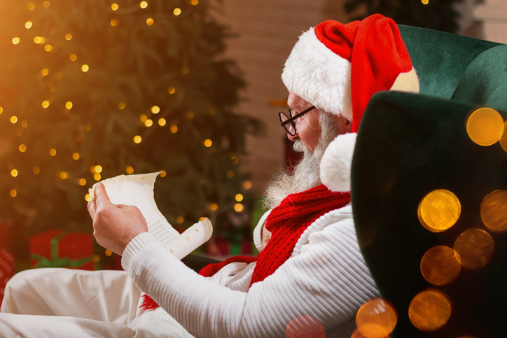 Serious Santa Claus sitting at his home and reading letters or wish list near the fireplace and Christmas tree with gifts. Image: iStock/monstArrr_