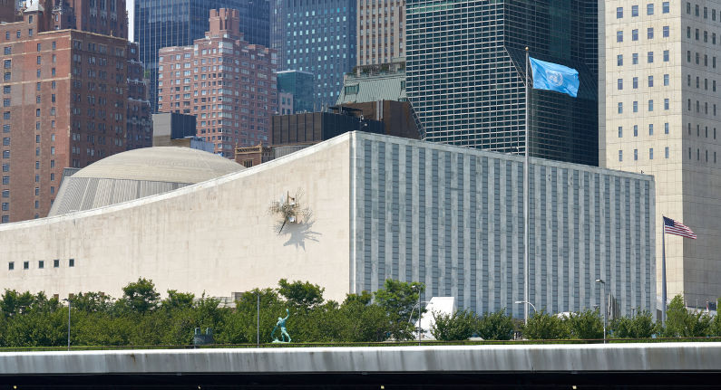 New York, The United Nations General Assembly Building at UN Headquarters in East Midtown Manhattan, NYC. The building is seen from the East River. The UN Flag flies next to the building and sculptures from the UN Collection can be seen.