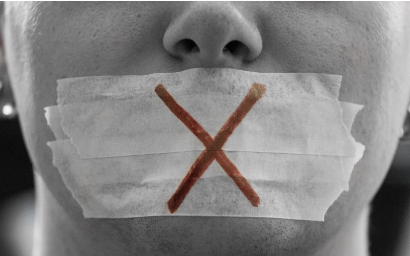 "A male face with a taped mouth and a red cross on it symbolizing censorship.Censorship is all around.Freedom of speech is the political right to communicate one's opinions and ideas via speech. Image: iStock / FL-photography