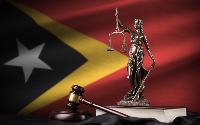 Timor Leste flag with statue of lady justice, constitution and judge hammer on black drapery. Concept of judgement and punishment. Timor Leste flag. Image: Contributor: Ingram Publishing / Alamy Stock Photo Image ID: 2S60EW9