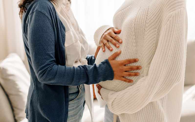 Young woman touching a surrogate mother's belly bump. Young woman spending time with her surrogate at home. Woman feeling the movement of a pregnant woman's baby. Image:iStock/JLco-Julia-Amaral