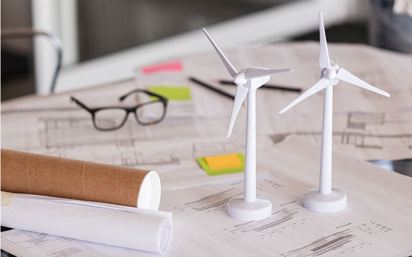 Close up of wind turbines on a table with architectural plan blueprints. Two models of wind turbines on desk with architecture layout and paper. Green and eco sustainable architecture. Image: iStock / Ridofranz