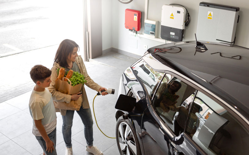 Son and mother plug EV charger from home charging station to electric vehicle. Home battery and clean energy now and future. Image iStock Basilico Studio Stock