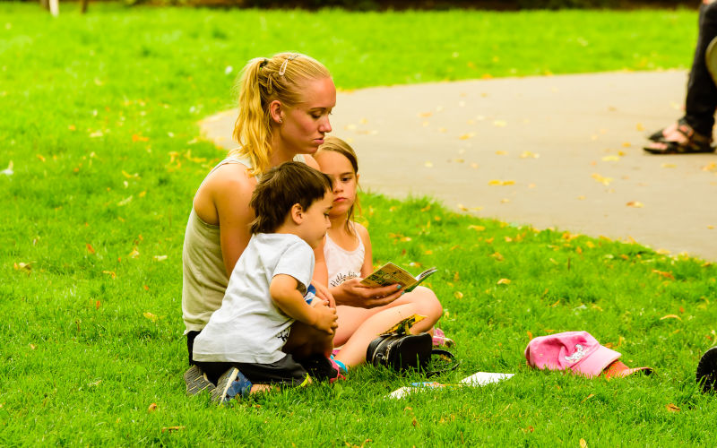 Young adult woman reading a small book for two children in a public park. Image iStock Reimphoto