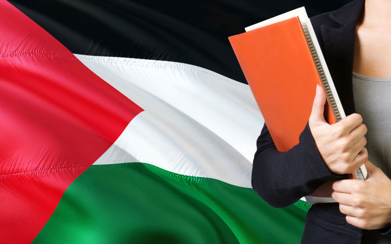 Palestinian education Young woman standing with the Palestine flag in the background. Teacher holding books, orange blank book cover. Image iStock sezer ozger