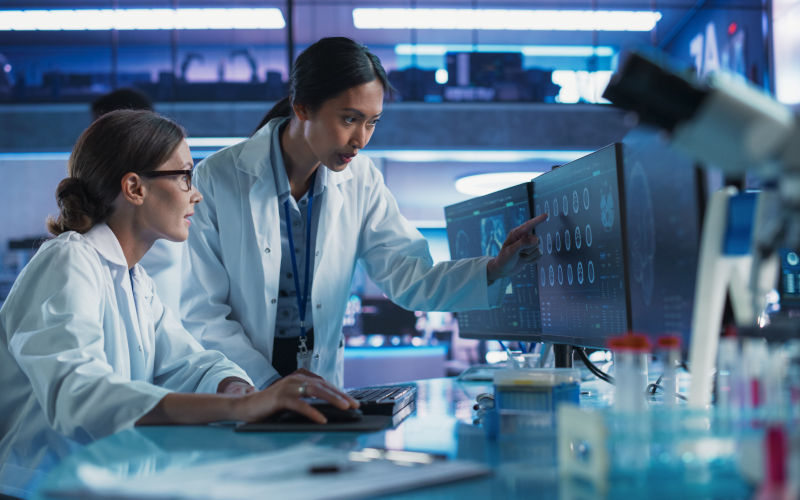 Women in STEM talking In Medical Research Center And Using Desktop Computer To Analyze MRI Scans Of Brain. Surgeon And Neuroscientist discussing solutions for treating cancer. Image iStock gorodenkoff