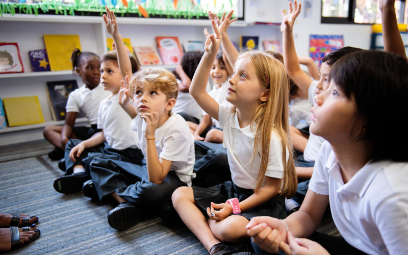 Diverse group of young students, boys and girls, sit on the floor in a classroom raising hands. Image iStock itRawpixel