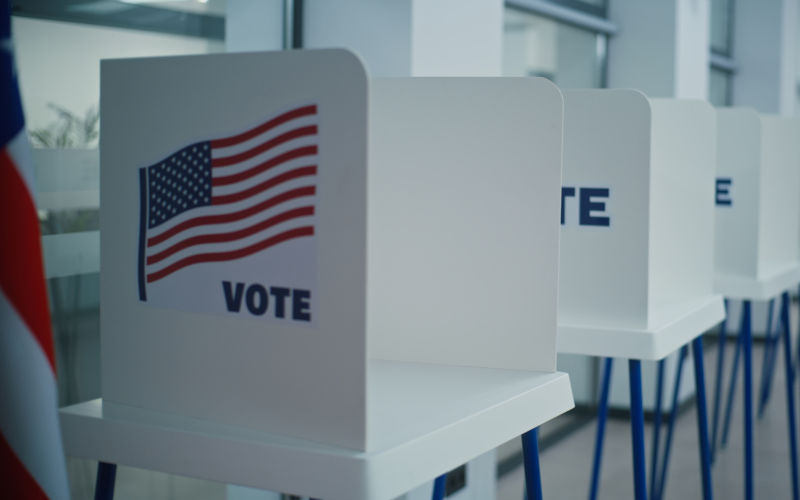 Voting booths with American flag logo in bright polling station office. Political races of US presidential candidates. Image iStock EvgeniyShkolenko