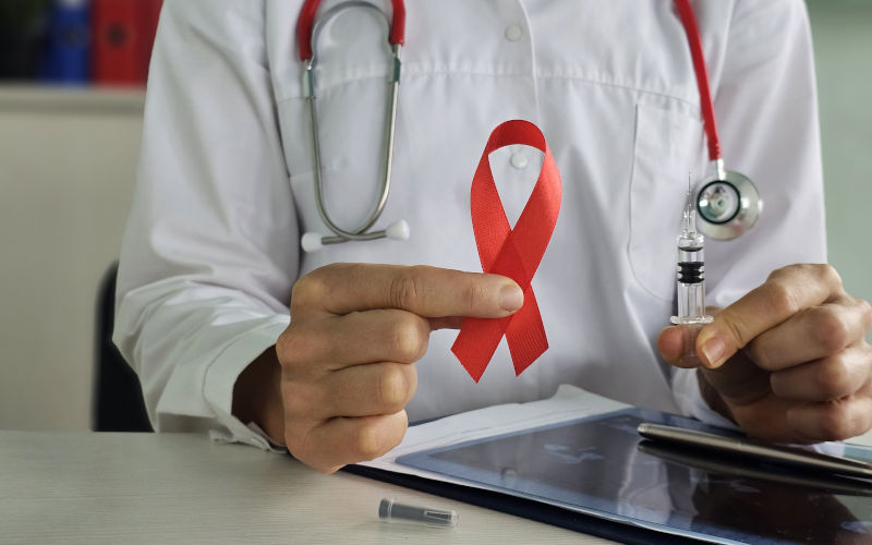 A health professional displays a red ribbon while holding a syringe, emphasizing the importance of HIV AIDS awareness and the vital role of vaccinations in healthcare. Image iStock Nadzeya Haroshka
