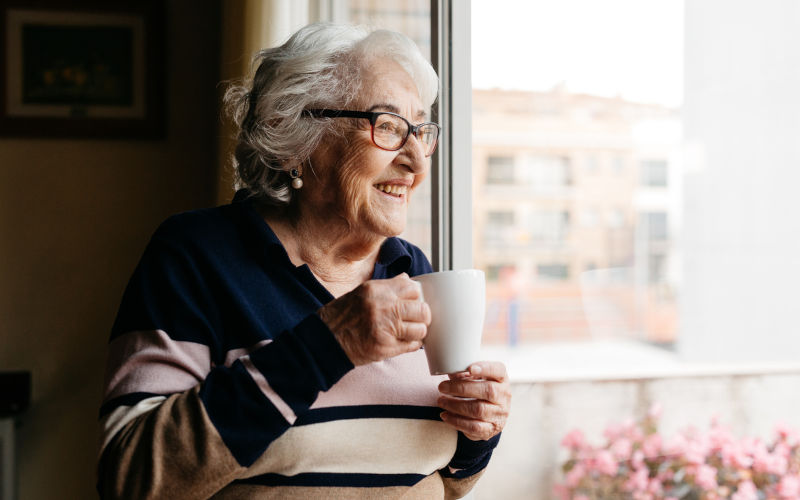 Elderly woman enjoys a warm cuppa while looking out the window. Image iStock Jordi Salas