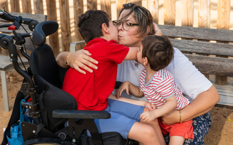 Mother showing affection to her sons, one boy with intellectual disability sitting in his wheelchair, creating a loving family moment. Image iStock Jordi Mora Igual