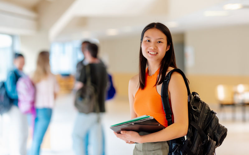 Portrait of an Asian student with a backpack and books in the hallways of a university campus with her classmates in the background. Image iStock Alberto Menendez
