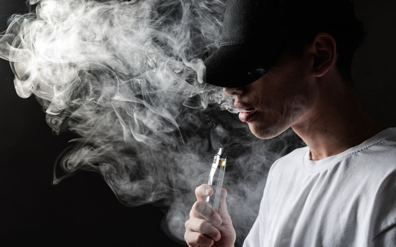 A young man with a cap exhales vapor from an ecigarette, highlighting the dangers of vaping and nicotine addiction in a moody atmosphere. Image iStock Sergii Kolesnikov