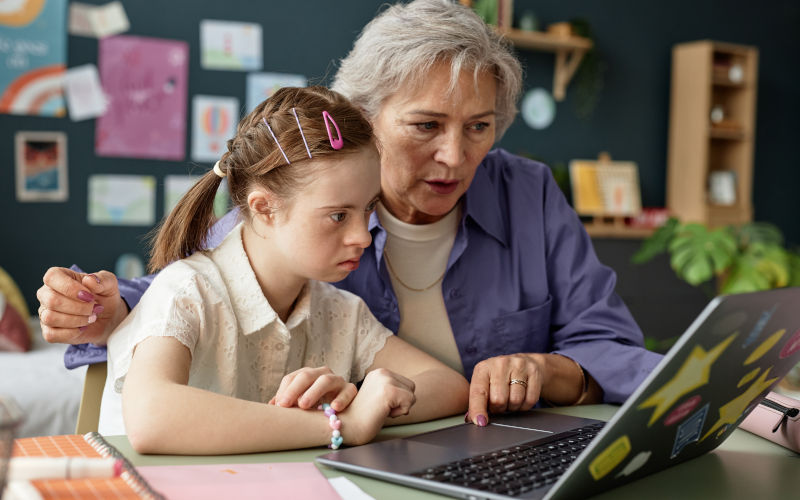 Senior woman guiding young girl with Down syndrome on laptop while sitting at a table in cozy home office, surrounded by educational materials. Image iStock SeventyFour