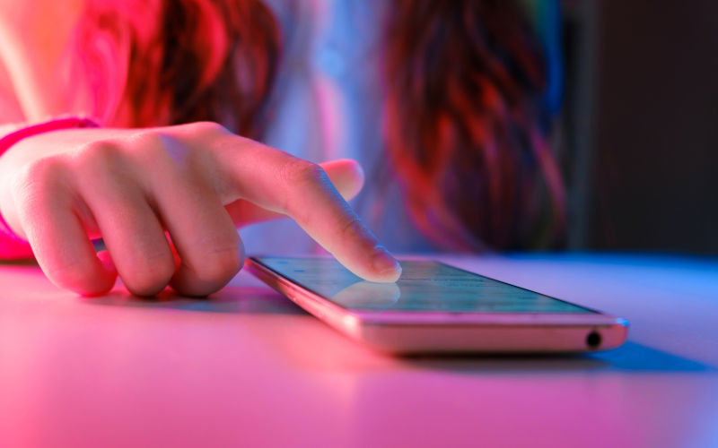 Hands of teen girl scrolling on phone in a pink lit room. Image iStock Marina Demidiuk