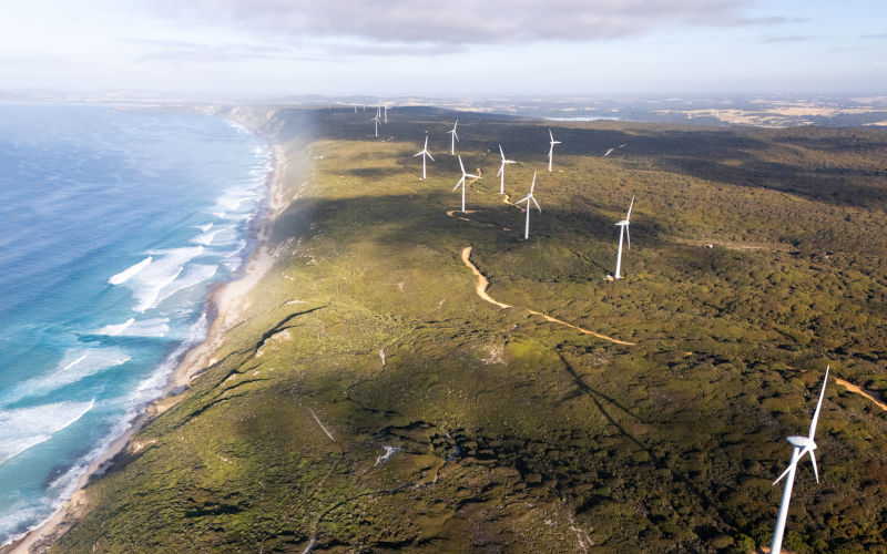Aerial view of wind turbines along a scenic coastline with ocean waves and green landscape. Clean energy infrastructure in a natural environment for renewable energy, sustainability, climate change, and ecofriendly power generation. Image iStock Mystockimages