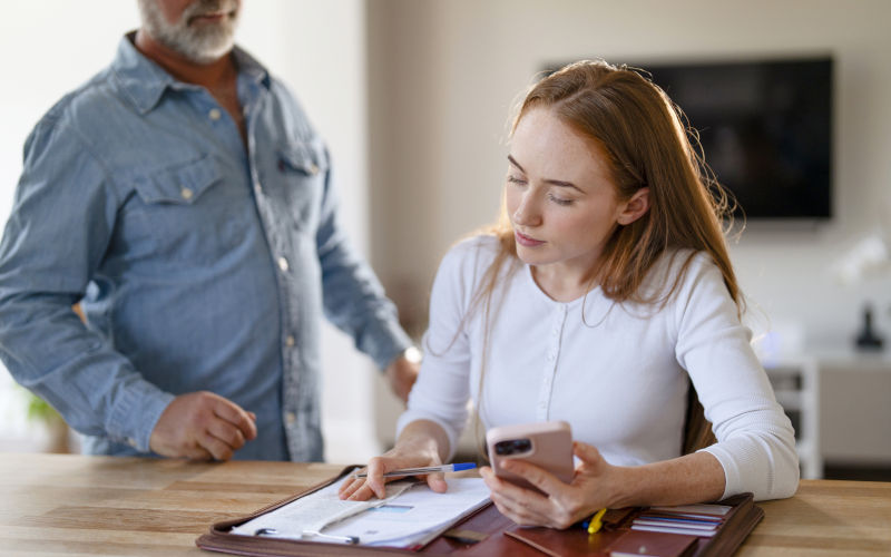 Young woman sits at a wooden table, looking at her phone and writing on a clipboard containing receipts and documents. An older man stands behind her, watching in a modern home. ImageiStock Iryna Melnyk