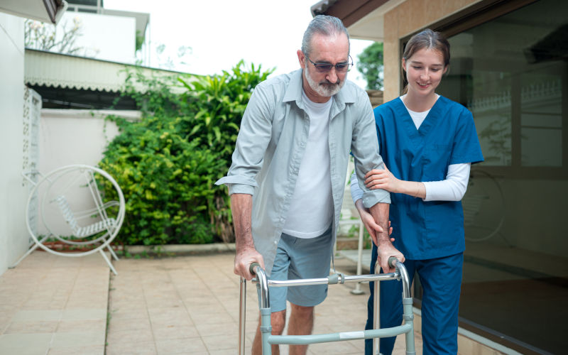 Elderly man holding a walking aid while being assisted by a young female caregiver learning to walk outside the home. Image iStock Sunan Wongsanga