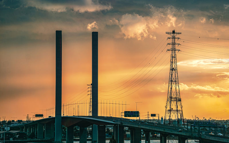 The twin concrete towers of Bolte Bridge rise beside an electricity transmission tower and power lines beneath sunlit clouds in Melbourne, Victoria, Australia. Image iStock AWWW