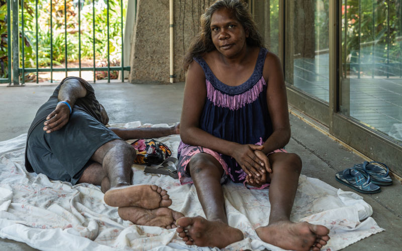 Darwin Australia February 22, 2019 Closeup of young aboriginal woman sitting in shade of building Sleeping man on her side. Image iStock ClaudineVM
